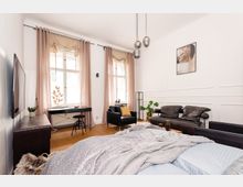 Helles, modernes Schlafzimmer mit großen Fenstern und lichtdurchlässigen Vorhängen, einem Bett im Vordergrund, einem Schreibtisch mit Stuhl sowie einer Sitzecke mit schwarzen Ledersesseln und Sofas. Die Wände sind weiß, mit dekorativen Leisten, und eine minimalistische Pendelleuchte hängt von der Decke.