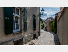 Schmale gepflasterte Gasse mit historischen Gebäuden aus Putz und Ziegeln, teilweise begrünt; links ein Haus mit Fensterläden und einer alten Straßenlaterne. Im Hintergrund sind Bäume und blauer Himmel mit Wolken sichtbar.
