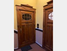 Zwei holzgeschnitzte Wohnungstüren in einem Treppenhaus mit gelb gestrichenen Wänden. Die Türen besitzen ovale Fenster mit Sprossen und dekorative Verzierungen, eine Klingel ist an der Wand montiert.