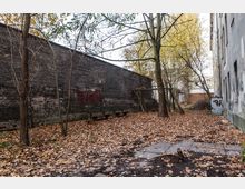 Ein leerer Hinterhof mit herbstlichem Laub bedeckt, umgeben von einer hohen Ziegelmauer und einem grauen Gebäude. Ein paar kahle Bäume und einfache Holzbänke prägen die Szenerie, während ein vernachlässigter Schaukelrahmen seitlich steht.