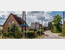 Straßenszene in einem Wohngebiet mit Einfamilienhäusern, die rote und graue Ziegeldächer haben. Links im Bild steht ein Straßenschild mit der Aufschrift „Rudolf-Breitscheid-Straße“, umgeben von Gärten, Bäumen und einem teilweise bewölkten Himmel.