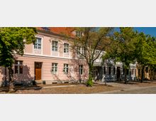 Straßenansicht einer Reihe historischer Wohnhäuser mit pastellfarbenen Fassaden, weißen Fensterrahmen und roten Ziegeldächern. Davor verläuft eine gepflasterte Straße mit Gehweg und mehreren großen Bäumen, die Schatten auf die Fassaden werfen.