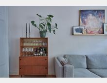 Holzkommode mit offenen Regalfächern, in denen Gläser und Spirituosen stehen, dekoriert mit zwei schwarzen Kerzenhaltern und einer grünen Zimmerpflanze auf der Oberseite. Daneben ein graues Sofa und eine minimalistische Bilderleiste mit mehreren Kunstwerken an einer weißen Wand.