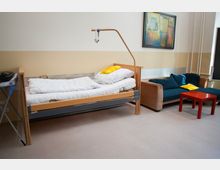 Zimmer mit einem Krankenhausbett aus Holz mit metallischem Rahmen, ausgestattet mit weißer Bettwäsche und gelbem Kissen. Daneben stehen ein Sofa mit blauen Kissen, ein roter Couchtisch und ein Wandbild im Hintergrund; die Wände sind beige und der Boden hell.