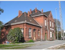 Backsteinbahnhofsgebäude mit einem Satteldach, mehreren Schornsteinen und weißen, bogenförmigen Fenstern. Der Bahnhof liegt an einer Gleisanlage und ist von Grünflächen umgeben. Ein Schild mit der Aufschrift „Zossen“ befindet sich an der Fassade.