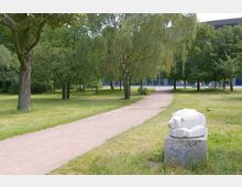 Ein Park mit einer grünen Wiese, mehreren Bäumen und einem geschwungenen Fußweg, der durch die Landschaft führt. Im Vordergrund steht eine weiße Skulptur eines Bären auf einem grauen Sockel; im Hintergrund ist ein modernes Gebäude zu sehen.