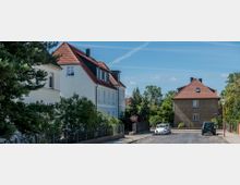 Wohnstraße mit freistehenden Einfamilienhäusern, die Satteldächer und helle Fassaden haben. Die Straße ist von Bäumen gesäumt, und am Straßenrand stehen geparkte Autos, darunter ein weißer Oldtimer.