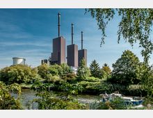 Industriegelände mit mehreren rechteckigen Gebäuden und hohen Schornsteinen, umgeben von grünem Laubwald. Im Vordergrund fließt ein Fluss, auf dem kleine Boote sichtbar sind.