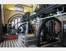 Industriehalle mit historischen Maschinen, darunter große schwarze Schwungräder und Rohre. Die Backsteinwände und Bodenfliesen im Schachbrettmuster prägen den Raum, ergänzt durch hohe Bogenfenster mit Tageslichteinfall.