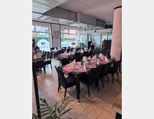 Ein Restaurant mit modernen, hellen Innenräumen, ausgestattet mit gedeckten Tischen, die rot-weiß-karierte Tischdecken und schwarze Stühle haben. Große Fensterfronten lassen Tageslicht herein und bieten einen Blick auf die Straße und die umliegenden Gebäude.