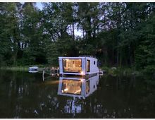 Ein modernes Hausboot mit großen Fenstern schwimmt auf einem ruhigen See, umgeben von dichtem Wald. Die Inneneinrichtung des Hausboots ist beleuchtet und spiegelt sich im Wasser wider.