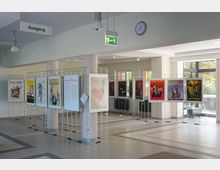 Ein moderner Innenraum mit einer Ausstellung von Postern, die auf Metallgittern montiert sind. Der Raum verfügt über große Fenster, durch die Tageslicht einfällt, sowie beschriftete Schilder wie „Ausgang“ und eine Wanduhr.