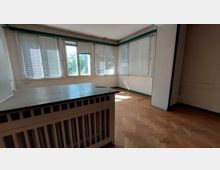 Ein leerer Raum mit hellem Parkettboden und mehreren Fenstern mit weißen Jalousien, die viel Tageslicht hereinlassen; im Vordergrund steht ein Heizkörper mit dekorativer Verkleidung.