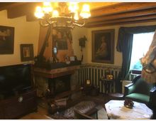 Gemütliches Wohnzimmer im klassischen Stil mit Holzbalkendecke, gemusterten Tapeten und einem großen gemauerten Kamin. Der Raum ist dekoriert mit antiken Möbeln, Gemälden, einem Kronleuchter und kleinen Sammlerstücken; ein Fenster mit schweren Vorhängen lässt diffuses Tageslicht herein.