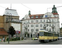 Straßenszene mit einer gelben Straßenbahn der Linie 62, die vor einem historischen, weiß verputzten Gebäude mit roten Dächern und dekorativen Fassadenelementen in Berlin-Köpenick vorbeifährt. Im Vordergrund sind Laternen, Werbetafeln und ein kleiner Baum zu sehen.