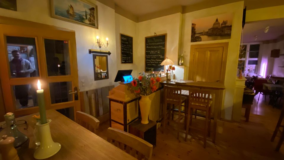 Gemütlicher Restaurantinnenraum mit Holztischen und -stühlen, einer Theke mit Barhockern sowie warmem Kerzen- und Lampenlicht. An den hellen Wänden hängen Bilder und Tafeln mit Speisekarte, daneben eine Holztür und ein Durchgang in einen weiteren Gastraum.