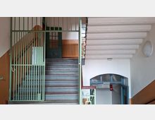 Treppenhaus in einem älteren Mehrfamilienhaus mit grauen Stufen, grünem Metallgeländer und einem Gittertor am Treppenaufgang. Sichtbar sind weiße Deckenbalken, ein bogenförmiger Durchgang mit blauem Türrahmen und zweifarbige Wände in Weiß und Beige.