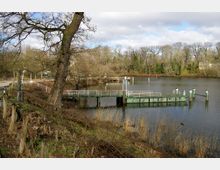 Eine Anlegestelle mit Metallgeländern und einem kleinen Bootssteg über einem ruhigen Fluss, umgeben von Bäumen ohne Laub. Im Vordergrund sind ein abfallendes Ufer mit kurzen Baumstümpfen und Gras zu sehen.