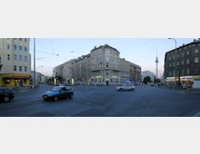 Straßenszene in einer Stadt mit einem großen Kreuzungsbereich, umgeben von mehrstöckigen Wohn- und Geschäftshäusern. Im Hintergrund ist der Berliner Fernsehturm deutlich sichtbar, während im Vordergrund Autos, Fahrräder und Fußgänger die Szene beleben.