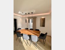 Esszimmer mit einem rechteckigen Holztisch, umgeben von acht grauen Polsterstühlen mit goldenen Details. An der Decke hängen vier schwarze, zylinderförmige Pendelleuchten; im Hintergrund befinden sich ein Fenster mit beigefarbenen Vorhängen und ein abstraktes Kunstwerk an der Wand.