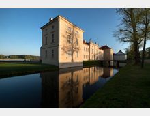 Ein helles, historisches Schloss mit mehreren Flügeln, reflektiert im Wasser eines umgebenden Kanals. Die Fassade ist beige, verziert mit hohen Fenstern und Statuen auf dem Dach; umgeben von Rasenflächen und Bäumen in einer parkähnlichen Landschaft.