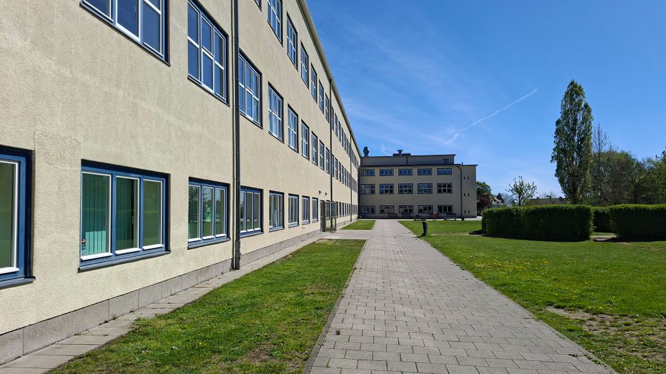 Langer, mehrstöckiger Gebäudekomplex mit heller Fassade und vielen rechteckigen Fenstern entlang eines gepflasterten Weges. Rechts liegen Rasenflächen mit geschnittenen Hecken und Bäumen, im Hintergrund steht ein weiteres Gebäude unter blauem Himmel.