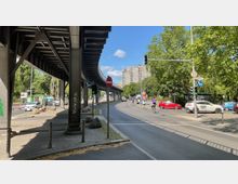 Straßenszene in einer Stadt mit einer erhöhten Bahntrasse aus Metall, die links im Bild verläuft. Rechts ist eine Straße mit mehreren Autos, Radfahrern und einer Ampel zu sehen, umgeben von Bäumen und in der Ferne ein mehrstöckiges Wohngebäude.
