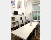 Ein kleiner Besprechungsraum mit einem langen weißen Tisch, umgeben von braunen Stühlen. An der Wand hängt ein Regal mit Ordnern und Büchern, darüber befinden sich gerahmte Poster; ein Fenster mit Gitter gibt Blick auf eine grüne Außenanlage frei.