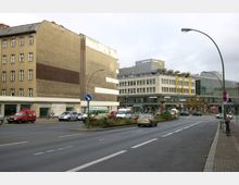 Straßenszene in einer Stadt mit einem mehrstöckigen, älteren Gebäude links und einem großen, modernen Kaufhaus von Karstadt rechts. Auf der Straße fahren Autos, und es sind geparkte Fahrzeuge sowie ein Fahrrad auf dem Gehweg zu sehen.