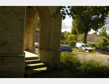 Blick durch einen gemauerten, gotischen Rundbogen mit Treppenstufen im Vordergrund auf eine ruhige, bewachsene Straße mit parkenden Autos und umliegenden Gebäuden. Die Szenerie ist von Bäumen und grünem Laub umrahmt.