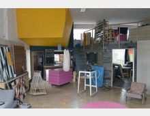 Ein kreativer Innenraum mit industriellem Charakter, gekennzeichnet durch eine gelbe Decke, eine Wandverkleidung aus Holzlatten und verschiedene Möbel, darunter ein pinkfarbener mobiler Tisch und ein Boxsack in der Mitte. Im Hintergrund ist ein Loft-Bereich mit einer Wendeltreppe und Regalen zu sehen, und der Raum enthält Arbeitsmaterialien und Kunstobjekte.