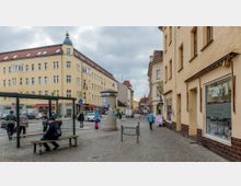 Städtischer Straßenzug mit gelb verputzten Wohn- und Geschäftshäusern, einem Wartehäuschen an einer Bushaltestelle, einer Litfaßsäule und einem breiten Gehweg. Im Hintergrund sind weitere Gebäude und ein schmaler Turm zu sehen, der Himmel ist bewölkt.