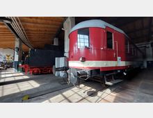 Ein historischer, rot-beiger Schienenbus steht in einer Lokhalle mit Holzdecke und Betonstützen. Im Hintergrund ist die Front eines schwarzen Dampflokomotiv-Tenders sichtbar.