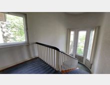 Treppenhaus in einem Gebäude mit weiß gestrichenen Wänden, einem schwarzen Holzhandlauf und weißen Geländerstreben. Im Hintergrund befinden sich große, gebogene Fenster mit Blick auf grüne Bäume.