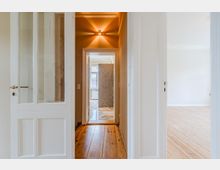 Blick durch einen schmalen Flur einer Wohnung mit weiß gestrichenen Türrahmen und Holzdielenboden, beleuchtet von einer Wandlampe an der Decke. Am Ende des Flurs ist ein Raum mit Fenster und einer gemusterten Fliesenwand zu sehen, rechts öffnet sich ein heller Raum mit Parkettboden.