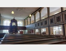 Innenansicht einer historischen Kirche mit hölzernen Kirchenbänken, einer reich verzierten Kanzel im Barockstil und einer umlaufenden Empore mit bemalten Wappen. Große Fenster lassen viel Tageslicht in den Raum, der von schlichten Hängeleuchten beleuchtet wird.