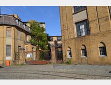 Ehemaliges Industriegebäude mit gelben Backsteinfassaden und großen Fenstern, umgeben von Kopfsteinpflaster. Im Vordergrund steht ein rostiges Metalltor mit einem Baum dahinter, das Gelände wirkt verlassen.