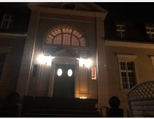 Nachtaufnahme eines größeren Gebäudes mit klassizistischer Fassade. Im Zentrum befindet sich ein beleuchteter Eingang mit einer dunklen Holztür, ovalen Fenstereinsätzen und darüber einem halbrunden Fenster; seitlich sind Kerzen platziert.