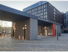 Eingangsbereich des Mercedes Platz in urbaner Umgebung mit grauem, modernem Flachdachgebäude. Im Vordergrund befinden sich ein Zugang zur Tiefgarage und Geldautomaten, dahinter ein mehrstöckiges Gebäude mit Hotelaufschrift "Hotel Indigo".