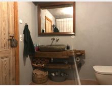 Rustikales Badezimmer mit einer Waschschale aus Stein auf einem Holzwaschtisch, darüber ein großer Holzrahmenspiegel. Neben der Waschgelegenheit befinden sich ein Handtuchhalter, ein Korb mit Badutensilien und im Vordergrund eine Toilette.