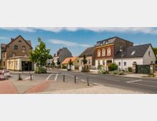 Straßenkreuzung in einer kleinen Ortschaft mit mehreren Wohnhäusern und einem Friseursalon in einem Eckgebäude. Die Umgebung ist von Gehwegen, Bäumen und Blumenbeeten gesäumt, und die Straße ist durch Zebrastreifen und Poller strukturiert.