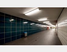 Unterführung mit weißen und dunkelgrünen gefliesten Wänden, beleuchtet von langen Deckenleuchten. Der Boden ist aus dunklem Material, und entlang des Gangs befinden sich Müllbehälter sowie vereinzelt Personen.