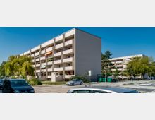 Wohnanlage mit mehreren vierstöckigen Gebäuden in moderner Bauweise, Balkonen und cremeweißen Fassaden. Im Vordergrund parkende Autos, Grünflächen, Bäume und ein gepflasterter Gehweg; blauer Himmel im Hintergrund.