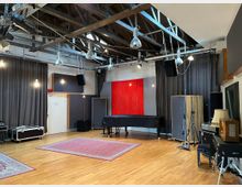 Großer Raum eines Tonstudios mit Holzboden, roten Teppichen und Vorhängen an den Wänden. Sichtbare Deckenkonstruktion aus Metall und Holz mit hängenden Lampen, ein Flügel in der Mitte sowie diverse Musikausrüstung wie Verstärker und Lautsprecher.