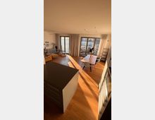 Wohnzimmer mit offenem Grundriss, Holzboden und großen Fensterfronten, die von bodenlangen Vorhängen eingerahmt sind. Im Raum stehen ein Esstisch mit Stühlen, eine Sitzecke mit Sessel und Dekor sowie eine moderne Kücheninsel im Vordergrund.