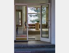 Geöffnete Glastüren führen in einen lichtdurchfluteten Raum mit großen Fenstern, Blick auf Bäume, einer modernen Stehlampe, einem runden Holztisch, einem Sessel und einer auffälligen Deckenleuchte. Davor befinden sich Stufen aus Stein.