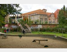 Spielplatz mit Sandfläche im Vordergrund, ausgestattet mit Wippen und einem Balancierbalken aus Holz. Im Hintergrund ein Backsteingebäude mit roten Ziegeldächern, daneben eine erhöhte Plattform mit Holzgeländer und Treppen, umgeben von Bäumen, Grafitti an einer Mauer und Rasen- und Sitzflächen.