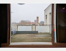 Blick durch eine geöffnete Balkontür auf eine Terrasse mit Holzboden und Glasgeländer. Im Hintergrund sind eine Kirchturmspitze, moderne Gebäude und kahle Bäume unter grauem Himmel zu sehen.