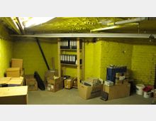 Ein Kellerraum mit gelb gestrichenen Backsteinwänden, Rohren an der Decke und verstreuten Kartons sowie Regalen mit Aktenordnern. Der Boden ist aus Beton, und der Raum ist spärlich beleuchtet.