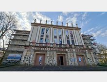 Frontansicht eines ehemaligen Lichtspieltheaters mit klassizistischer Fassade, hohen schmalen Fensterfeldern und Balustrade; über dem Eingang steht „LICHTSPIELTHEATER DER JUGEND“. Das Gebäude ist teilweise eingerüstet, die Eingänge sind verschlossen bzw. geöffnet, und die Außenwände zeigen Graffiti; davor verläuft eine breite, leere Vorfläche unter wolkigem Himmel.
